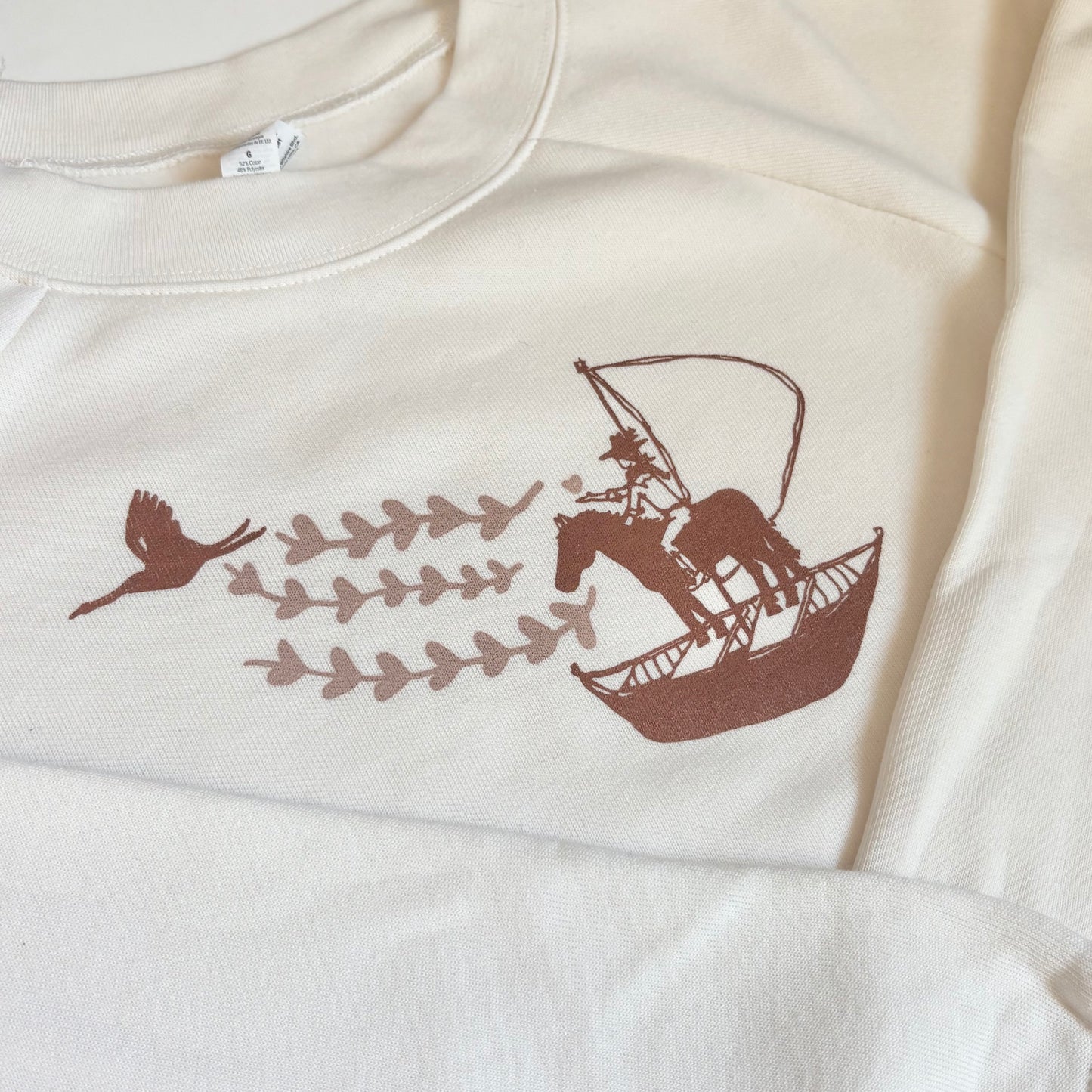 White sweatshirt with brown graphic design of a horse on a boat with birds and leaves.