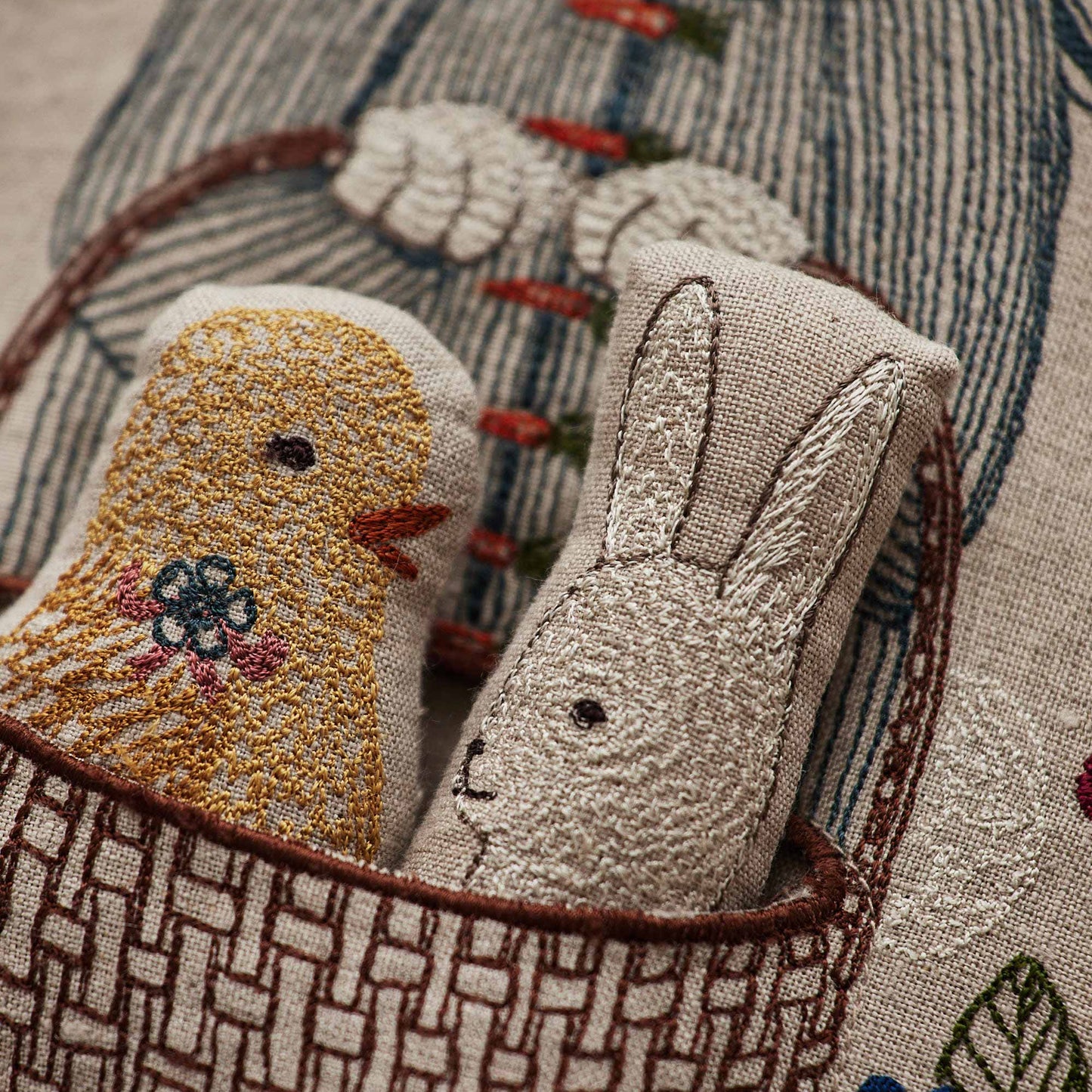 Coral and Tusk’s “Easter Bunny” embroidered linen pocket pillow featuring a festive rabbit carrying a basket with a chick and bunny inside, measures 12“ x 16”