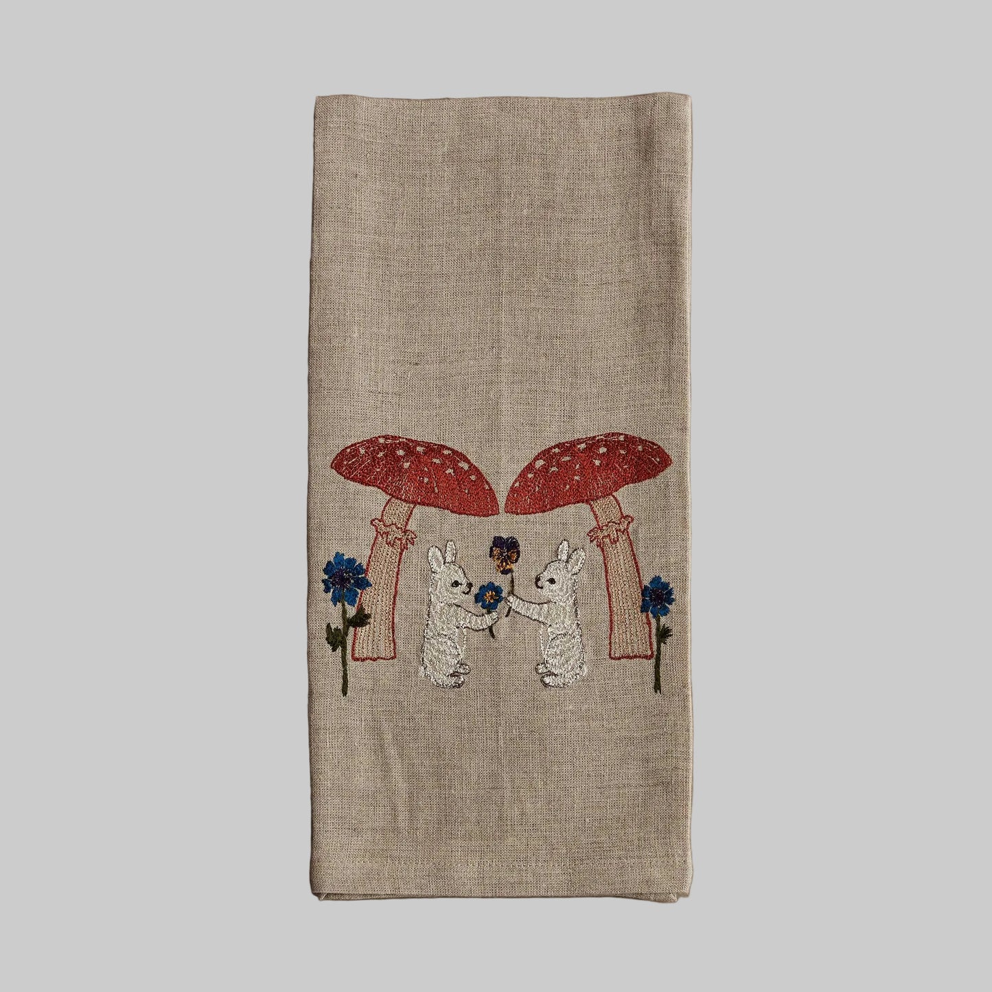 Coral and Tusk’s “Mushroom Bunny Friends” embroidered linen tea towel featuring two bunnies exchanging flowers under large mushrooms