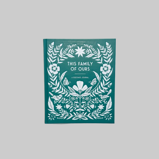 The cover of a beautiful keepsake journal titled "This Family of Ours". The cover is a teal color, embellished with intricate white papercut designs that include floral motifs and other decorative elements.