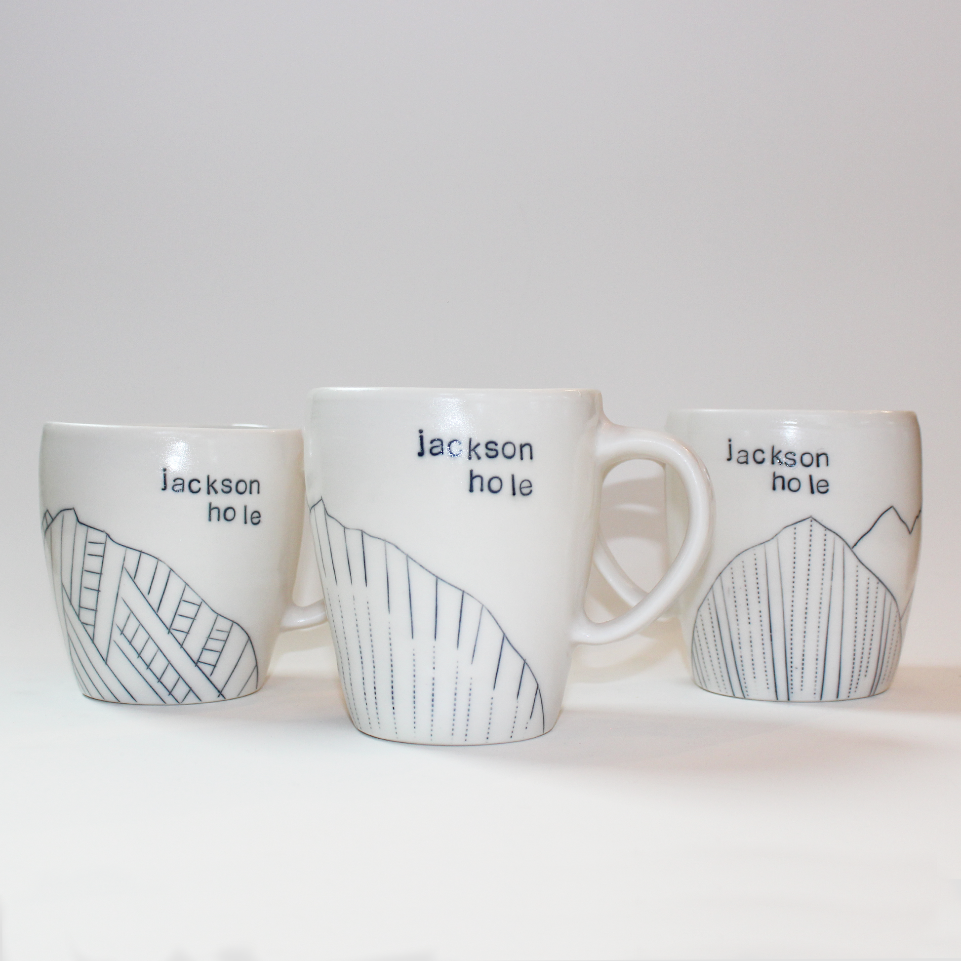 Three hand-thrown glazed porcelain mugs with 'Jackson Hole' text and mountain designs on them, made by Jenny Dowd