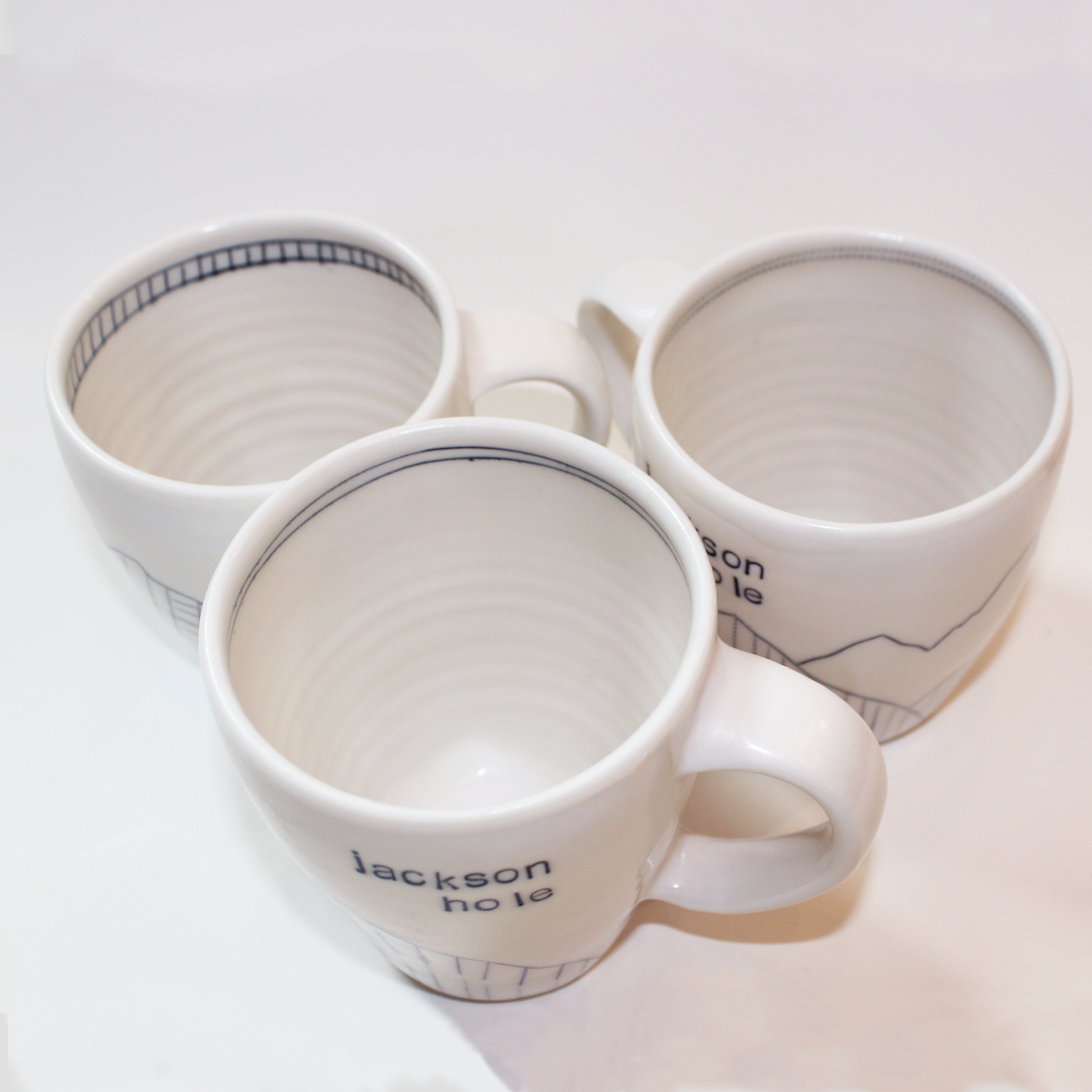 Three hand-thrown glazed porcelain mugs with 'Jackson Hole' text and mountain designs on them, made by Jenny Dowd