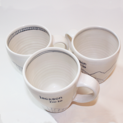 Three hand-thrown glazed porcelain mugs with 'Jackson Hole' text and mountain designs on them, made by Jenny Dowd