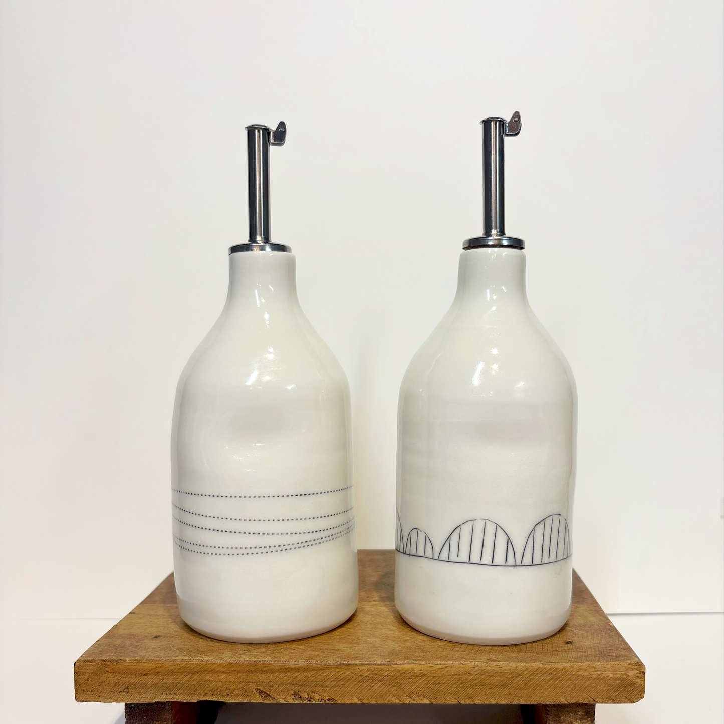 Two white ceramic oil cruet bottles with black line designs and silver nozzles 