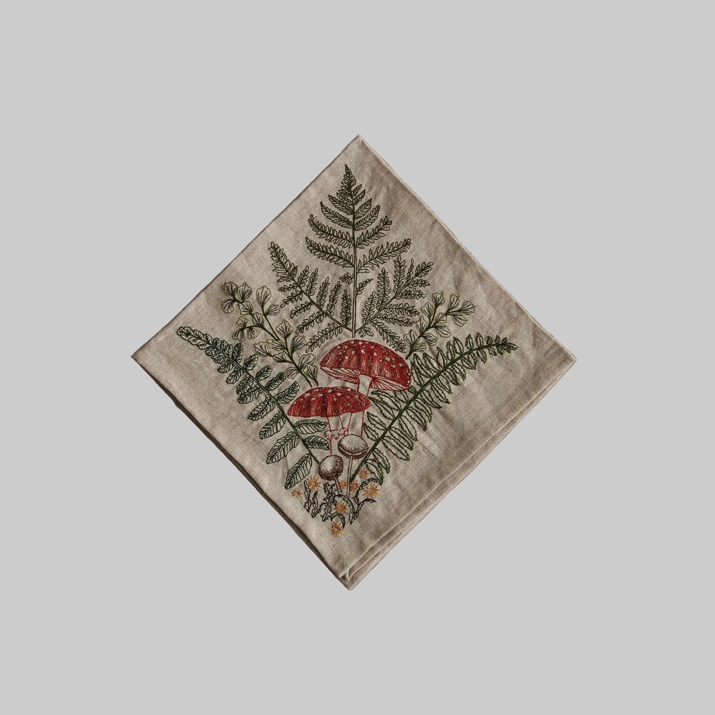 Coral and Tusk’s “Mushrooms and Ferns” embroidered linen dinner napkin featuring a bouquet of orange and brown mushrooms with fern fronds