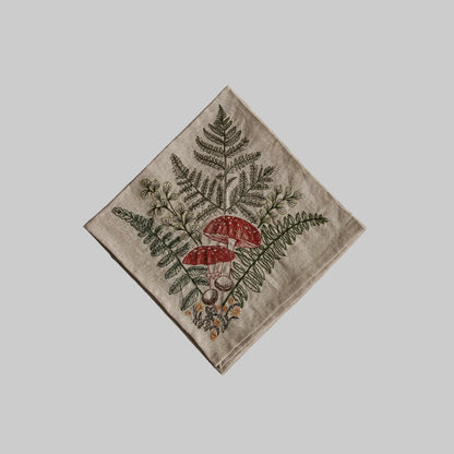 Coral and Tusk’s “Mushrooms and Ferns” embroidered linen dinner napkin featuring a bouquet of orange and brown mushrooms with fern fronds