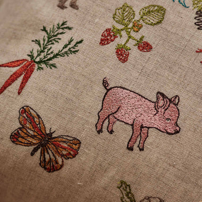 Coral and Tusk’s “Garden Gathering” embroidered linen pillow featuring colorful farm animals among pollinators, fruits, and vegetables, measures 12” x 16”