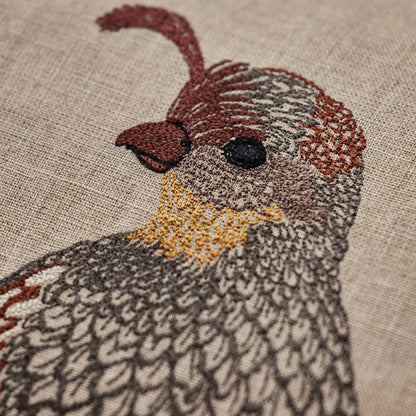 Coral and Tusk’s “Desert Quail Love” embroidered linen pillow featuring a pair of quails with gold, brown, and gray feathers, measures 12” x 16”