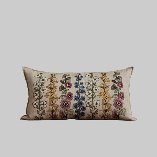 Coral and Tusk’s “Spring Stems” embroidered linen pillow featuring intricate delphiniums, dogwoods, forsythia, and hollyhocks, measures 14” x 26”