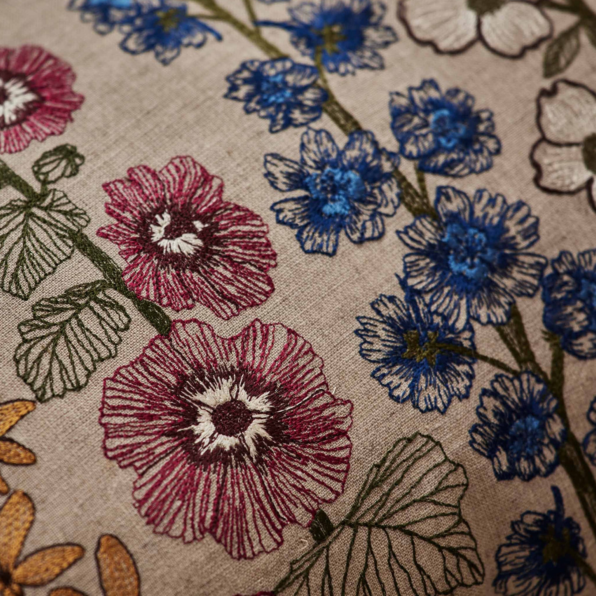 Coral and Tusk’s “Spring Stems” embroidered linen pillow featuring intricate delphiniums, dogwoods, forsythia, and hollyhocks, measures 14” x 26”