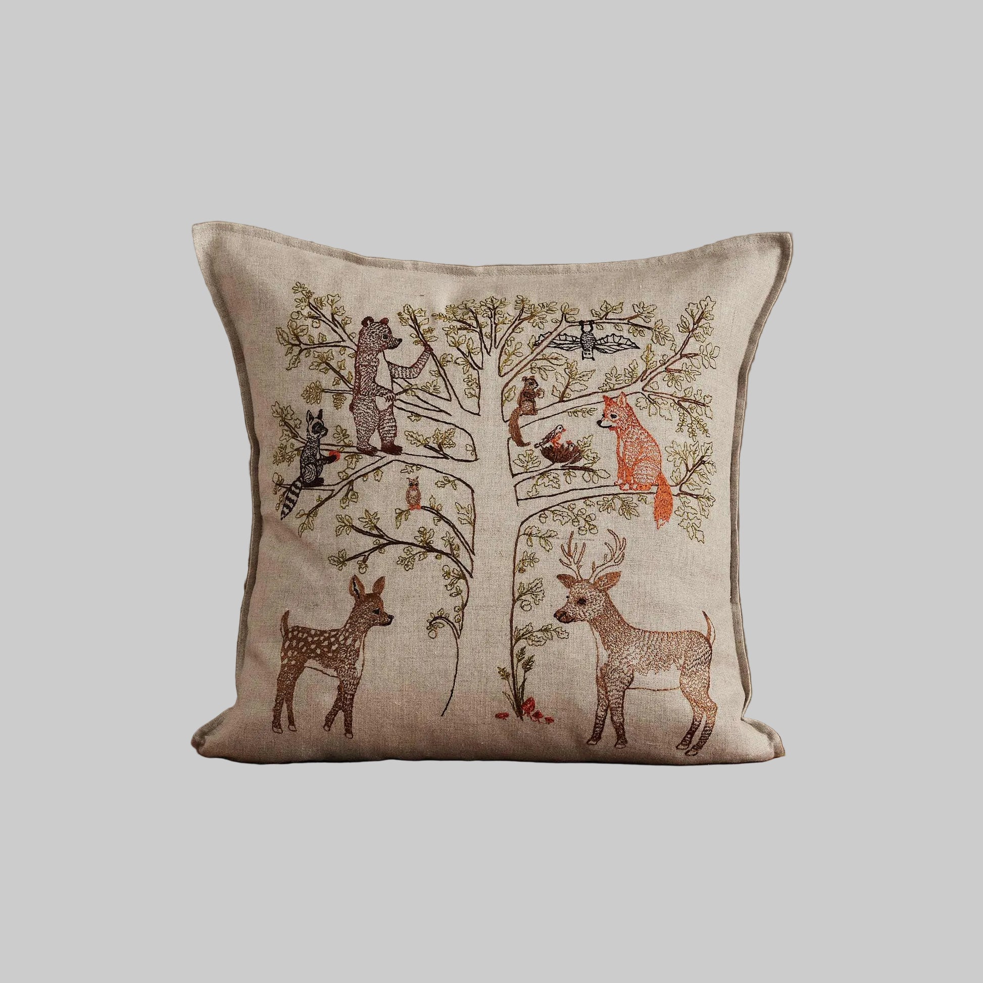 Coral and Tusk’s “Woodland Living Tree” embroidered linen pillow featuring a bear, raccoon, bird, fox, and deer living among towering trees and tiny mushrooms, measures 16” x 16”