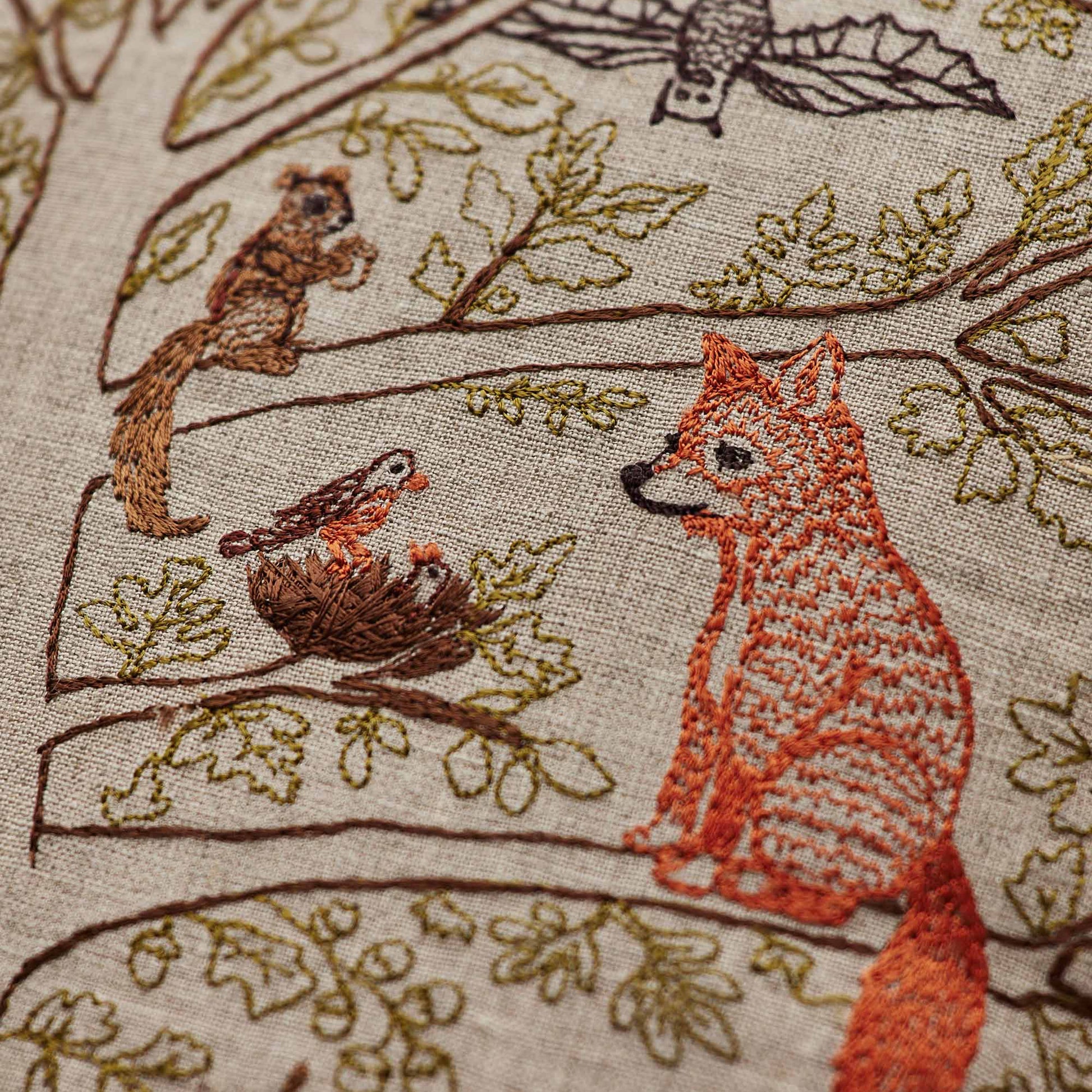 Coral and Tusk’s “Woodland Living Tree” embroidered linen pillow featuring a bear, raccoon, bird, fox, and deer living among towering trees and tiny mushrooms, measures 16” x 16”