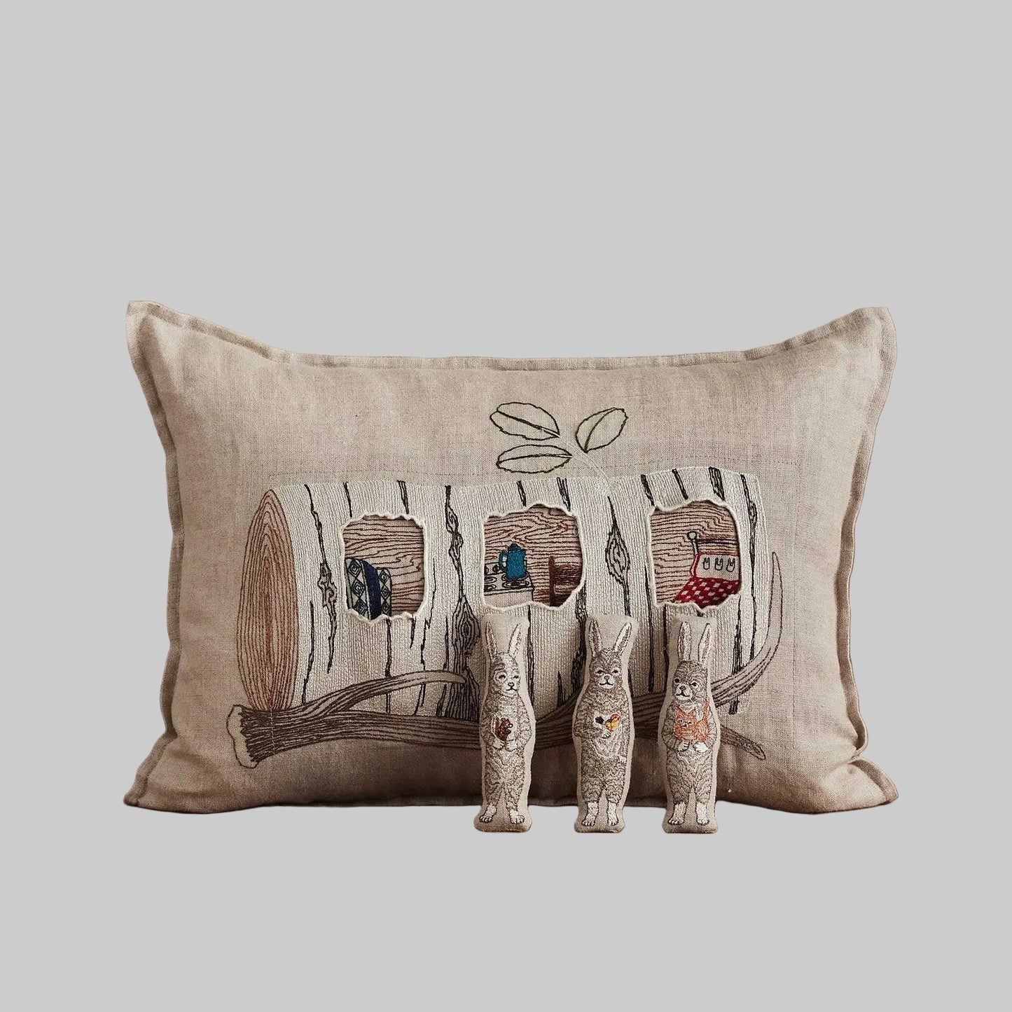 Coral and Tusk’s “Aspen Log Bunnies” embroidered linen pocket pillow featuring a family of three rabbits hiding away in their cozy tree stump home, measures 12” x 16”