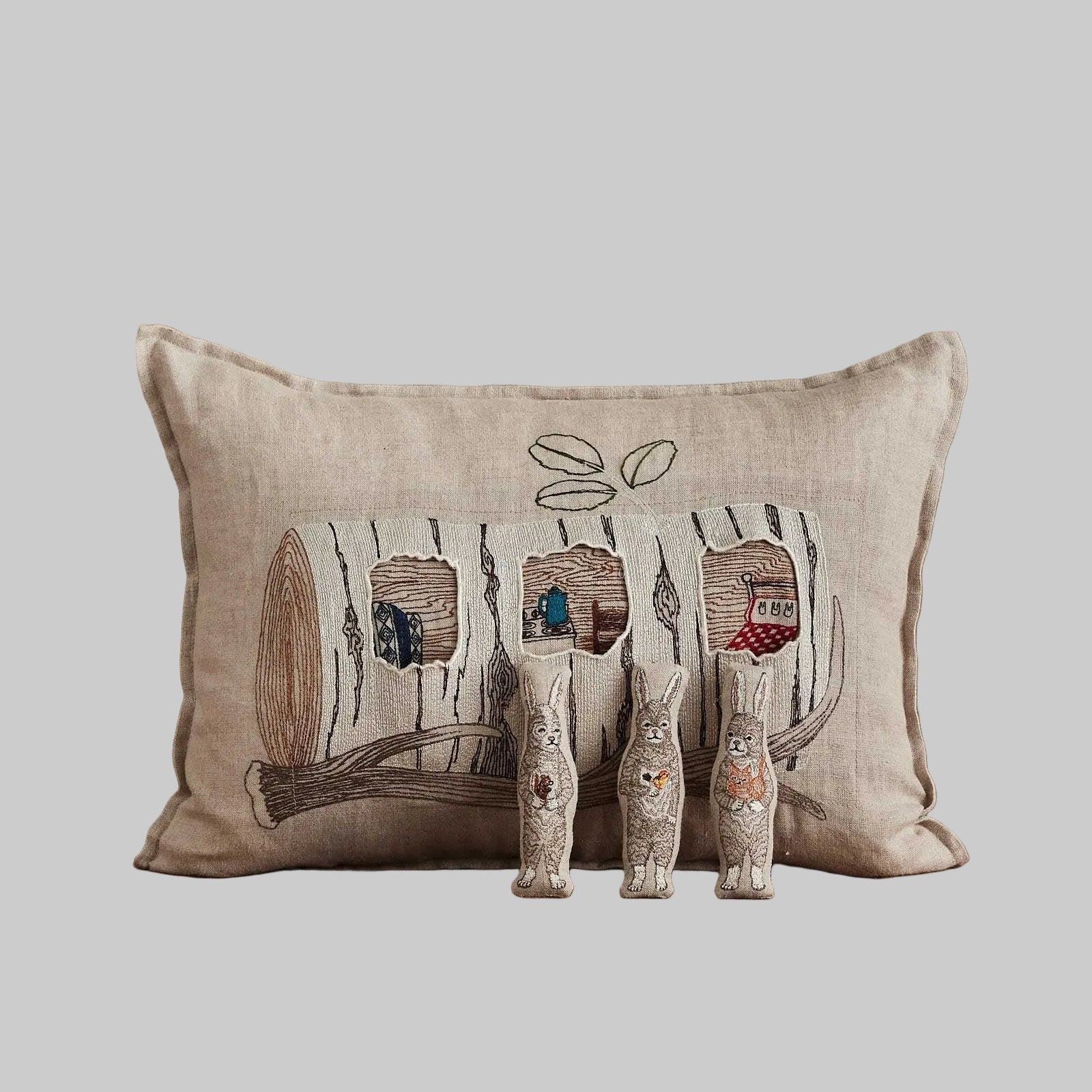 Coral and Tusk’s “Aspen Log Bunnies” embroidered linen pocket pillow featuring a family of three rabbits hiding away in their cozy tree stump home, measures 12” x 16”