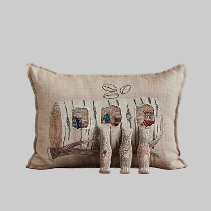 Coral and Tusk’s “Aspen Log Bunnies” embroidered linen pocket pillow featuring a family of three rabbits hiding away in their cozy tree stump home, measures 12” x 16”