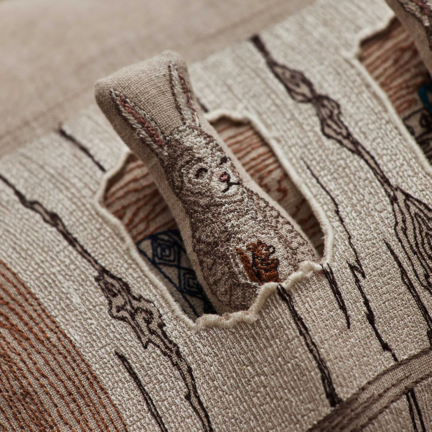 Coral and Tusk’s “Aspen Log Bunnies” embroidered linen pocket pillow featuring a family of three rabbits hiding away in their cozy tree stump home, measures 12” x 16”