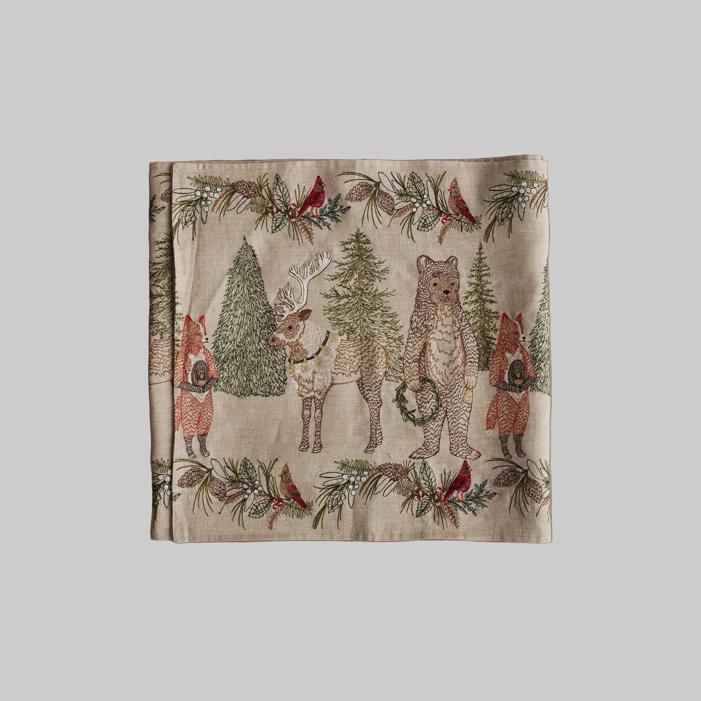 Coral and Tusk’s “Christmas Cheer” embroidered linen table runner featuring woodland animals holding wreaths surrounded by festive greenery