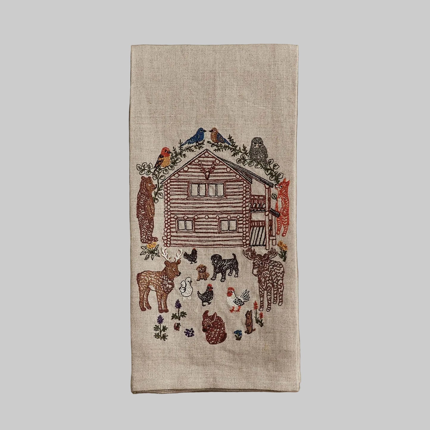 Coral and Tusk’s “Cabin Life” embroidered linen tea towel featuring a woodsy cabin surrounded by woodland creatures