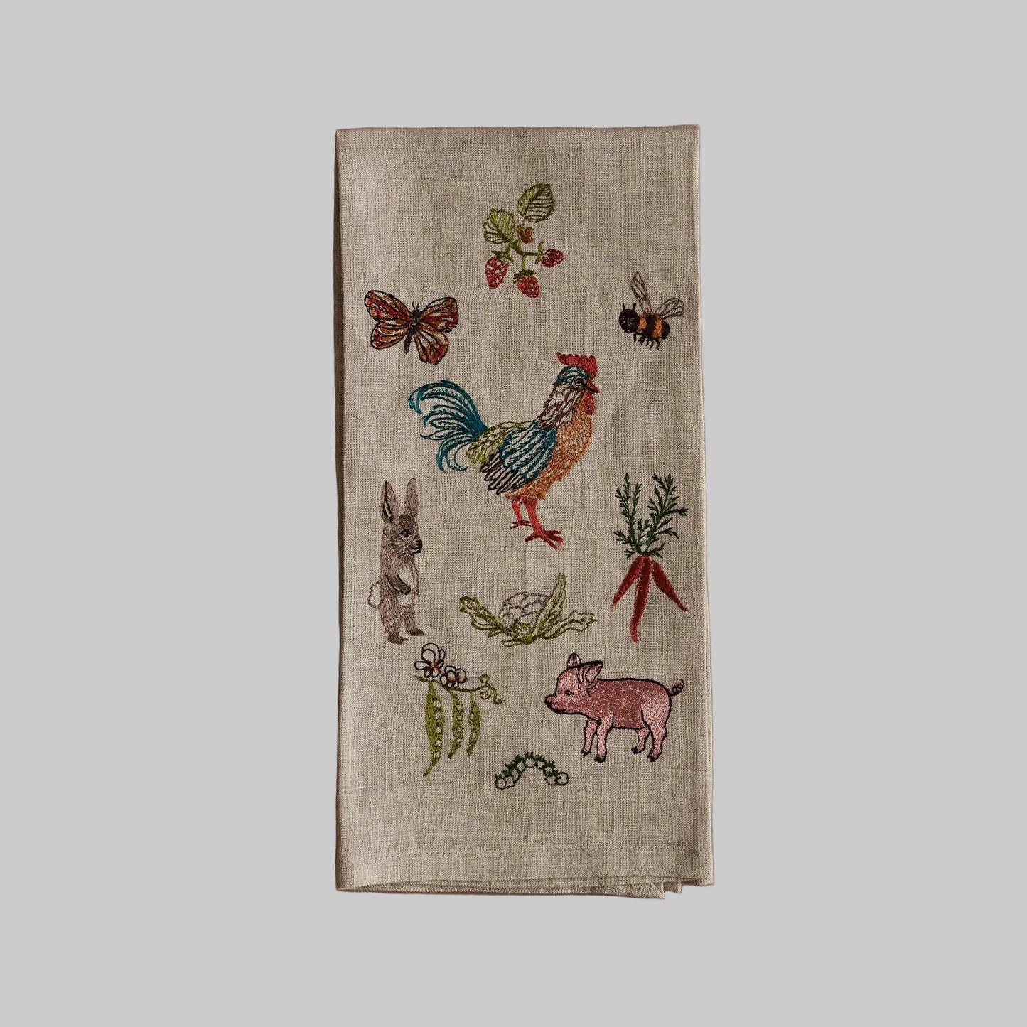 Coral and Tusk’s “Garden Gathering” embroidered linen tea towel featuring a group of farm animals, pollinators, and produce