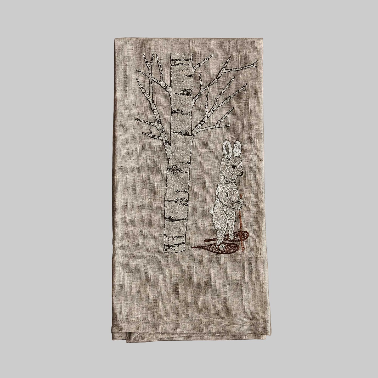 Coral and Tusk’s “Snowshoe Hare” embroidered linen tea towel featuring a white hair snowshoeing past an aspen tree