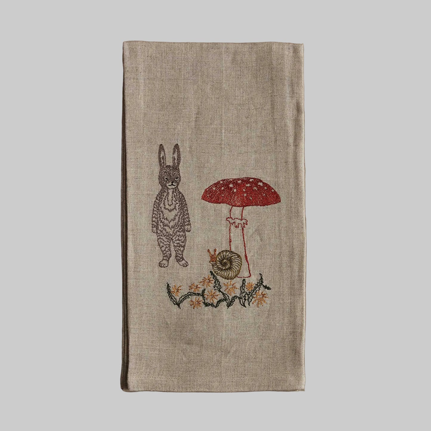 Coral and Tusk’s “Bunny with Mushrooms” embroidered linen tea towel featuring a gray bunny standing next to a tall mushroom