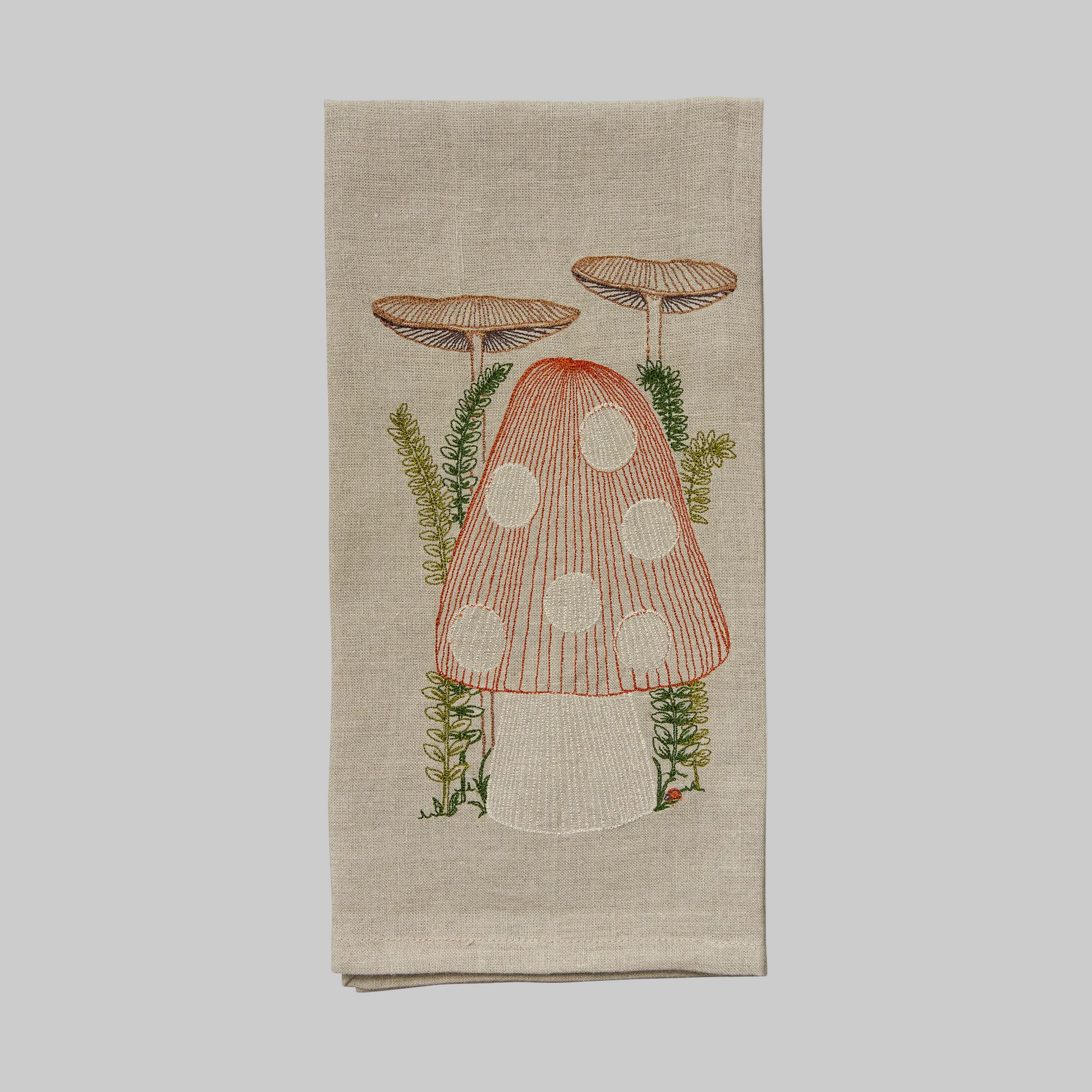 Coral and Tusk’s “Mushroom” embroidered linen tea towel featuring a large mushroom nestled in green sprigs