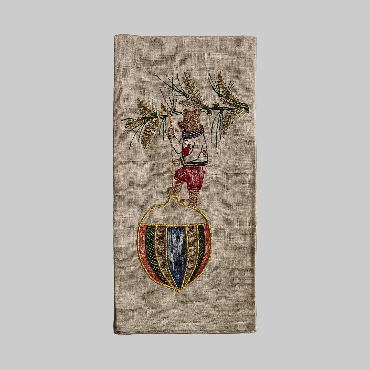 Coral and Tusk’s “Deck the Halls Bear” embroidered linen tea towel featuring a bear standing on a large Christmas ornament 