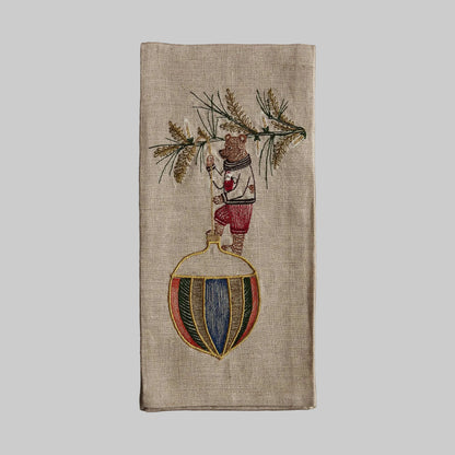 Coral and Tusk’s “Deck the Halls Bear” embroidered linen tea towel featuring a bear standing on a large Christmas ornament 
