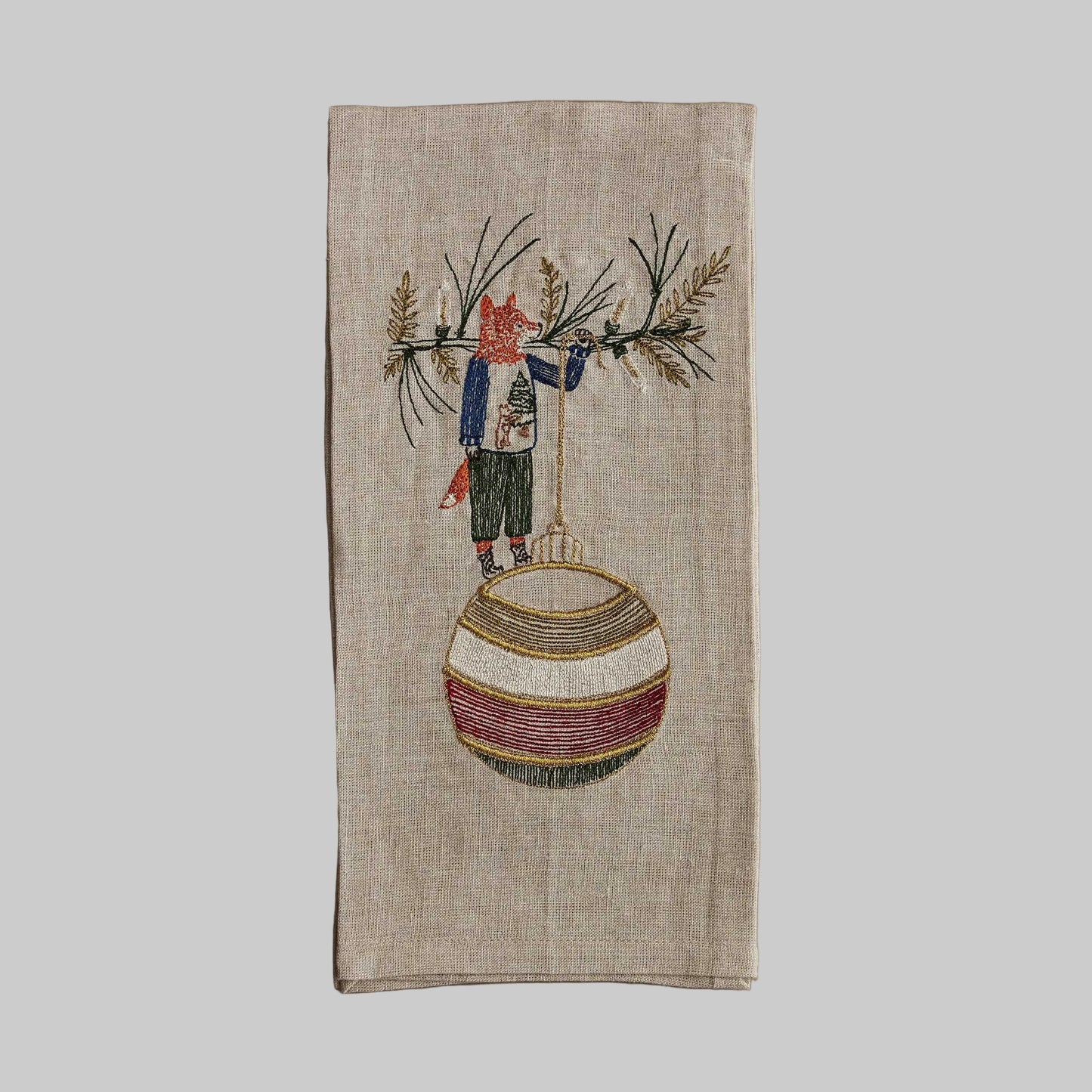 Coral and Tusk’s “Deck the Halls Fox” embroidered linen tea towel featuring a fox standing on a large Christmas ornament