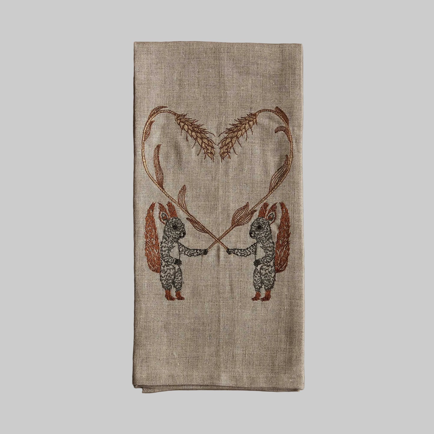 Coral and Tusk’s “Squirrels with Wheat” embroidered linen tea towel featuring two squirrels holding strands of wheat in the shape of a hearts