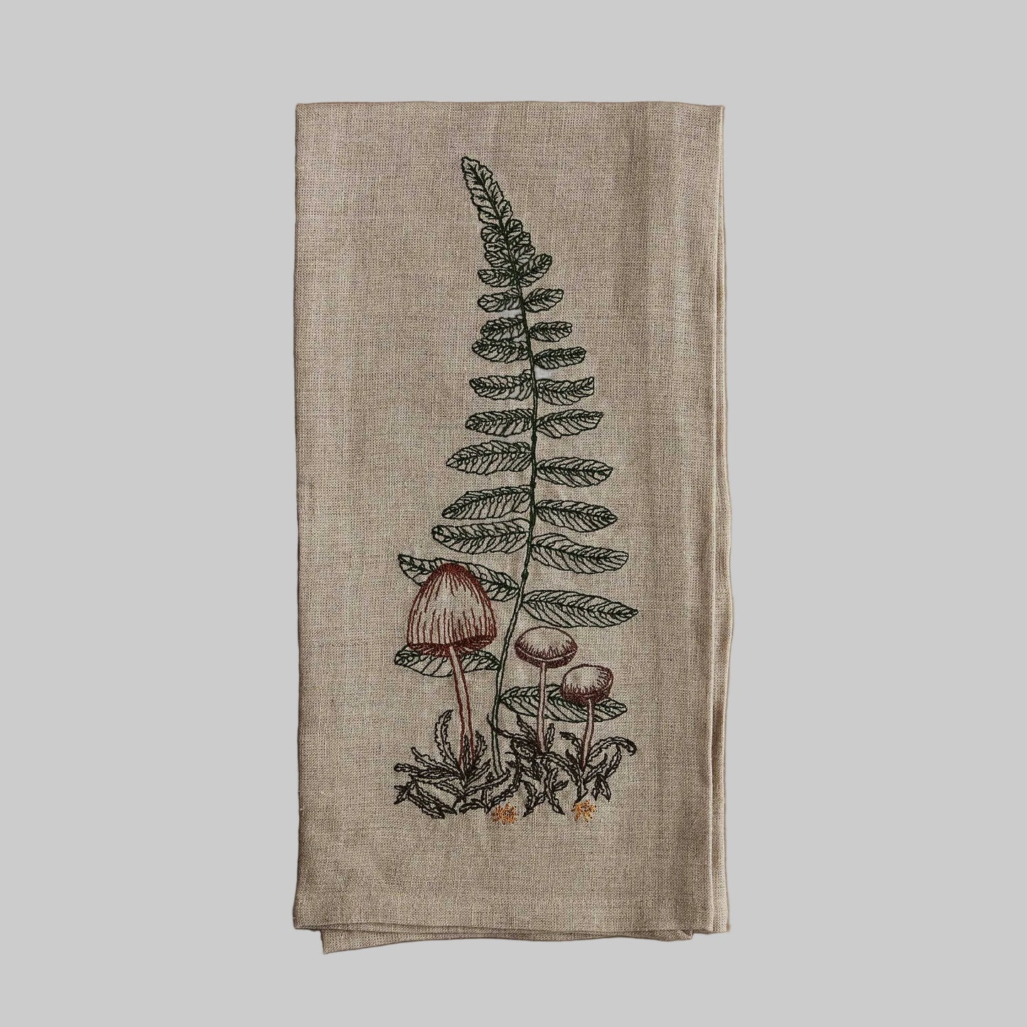 Coral and Tusk’s “Sword Fern” embroidered linen tea towel featuring small brown mushrooms in front of a fern frond