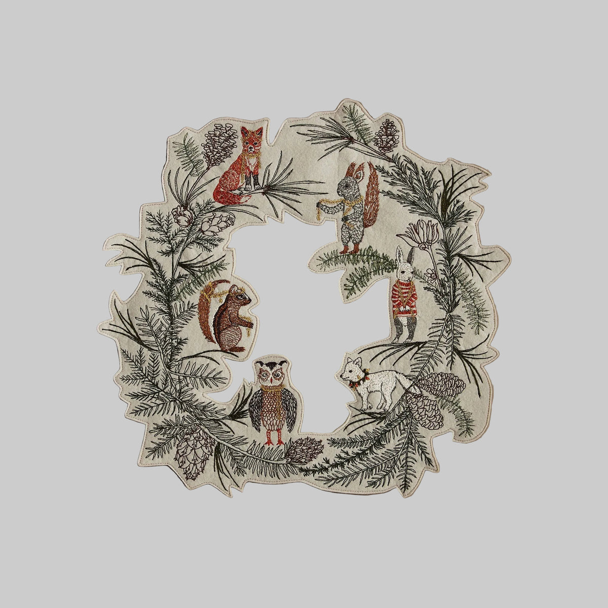 Coral and Tusk's "Tree Trimmers" embroidered holiday wreath featuring intricate pine needle designs and animal friends